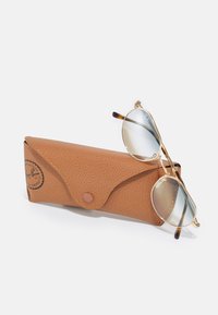 Occhiali da sole Ray-Ban con lenti blu, montatura in metallo dorato e dettagli in tartaruga, posizionati accanto a una custodia in pelle marrone con texture, dotata di chiusura a bottone.