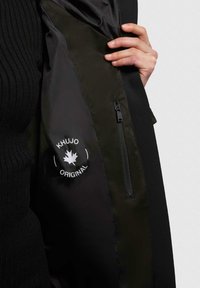 Sort jakke indvendig med glat, mørkegrøn foring; har en synlig lynlås lomme og et hvidt logo med et ahornblad-design.