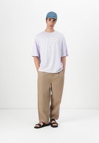 Světle fialové oversize tričko s bílým textem, béžové široké kalhoty a černé sandály; volnější džínová čepice dokončuje outfit.
