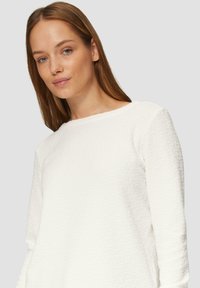 Haut blanc texturé à manches longues avec un col rond et un motif en tissu réhaussé doux. Design simple et minimaliste sans matériel visible.