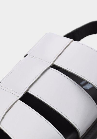 Λευκό δερμάτινο σανδάλι με πλατιές επικαλυπτόμενες λουρίδες και εμφανείς λευκές ραφές σε μαύρη σόλα πάνω σε γκρι φόντο.