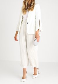 Blazer blanco con puños abiertos, top rosa claro, pantalones de pierna ancha a rayas beige, tacones blancos y un bolso de mano plateado.