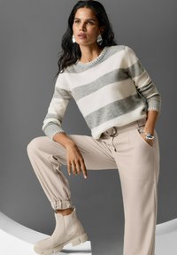 Frau posiert vor grauem Hintergrund, trägt einen grau-weißen gestreifen Pullover, beiges hoch tailliertes Hosen und beige klobige Stiefel sowie silbernen Schmuck.