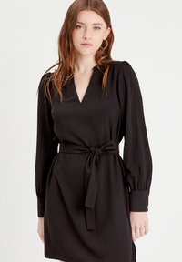 Robe noire avec un décolleté en V, des manches longues à volants, et une ceinture nouée à la taille. Caractéristiques d'un tissu texturé et de boutons décoratifs sur les poignets.