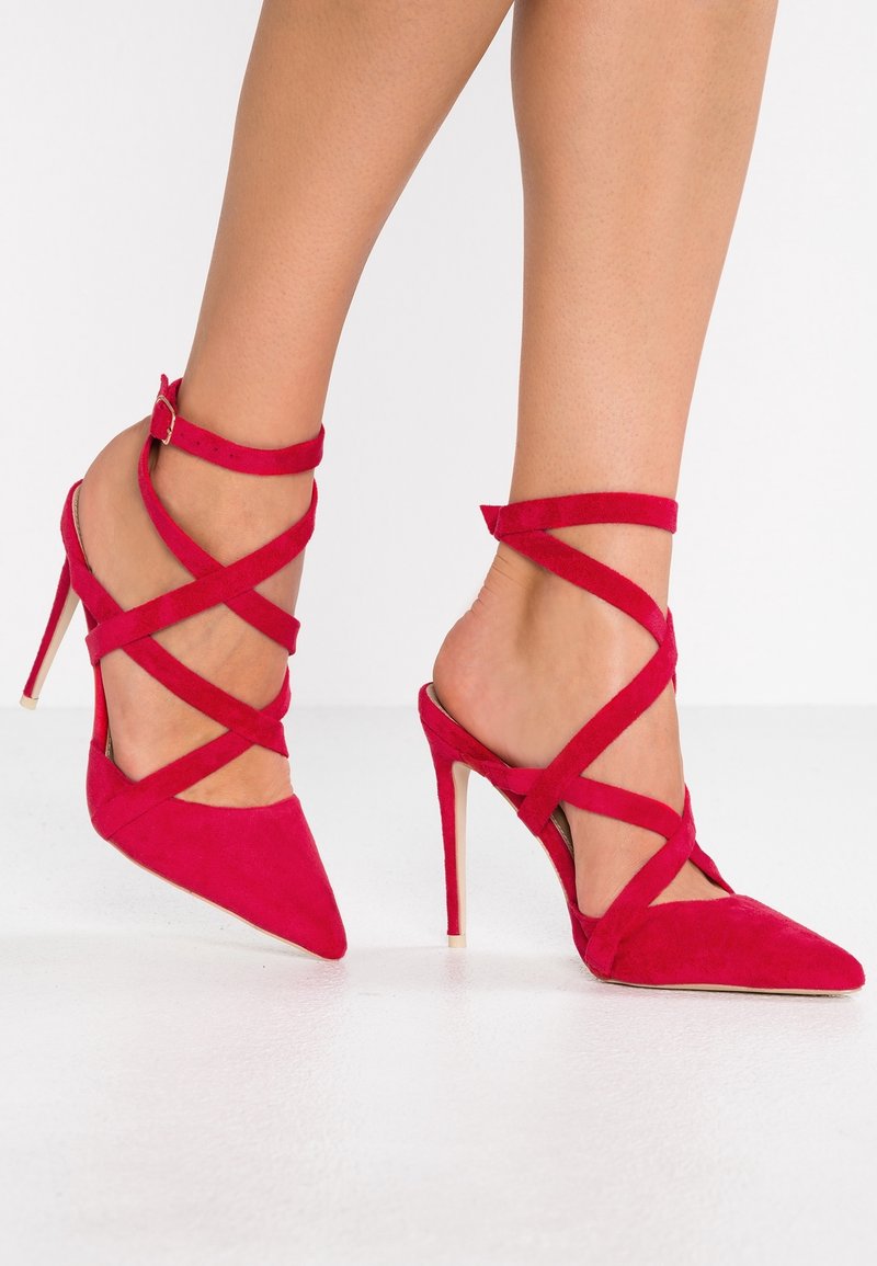 Jambes portant des talons aiguilles rouge vif avec plusieurs brides croisées à la cheville, debout sur un sol blanc.