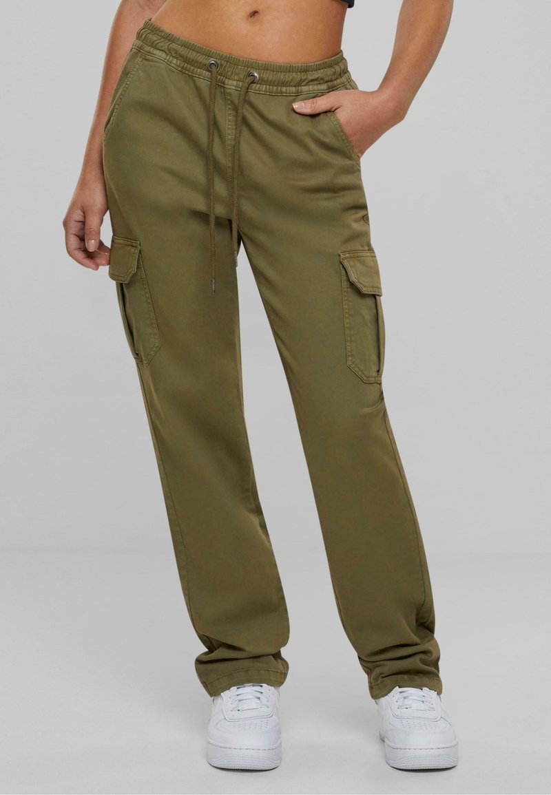 Partie inférieure du corps portant un pantalon cargo vert olive avec taille élastique et cordon, associé à des baskets blanches, debout sur un fond gris.