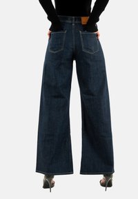 Jeans de pierna ancha de color azul oscuro, hechos de mezclilla, con cintura alta y grandes bolsillos traseros, con costuras en contraste y una parche de cuero marrón.