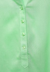 Grön skjorta med knäppning framtill, tillverkad av ett mjukt tyg, med krage, sex knappar och en liten ficka med en spetsig klaff.