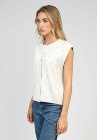 Cardigan sans manches en crème avec de petits motifs floraux brodés, col rond et fermeture à boutons sur le devant. Texture en tricot doux.