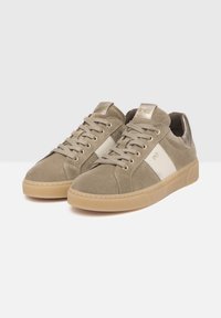 Baskets en daim beige avec une bande en cuir blanc, des accents métalliques sur le talon et une semelle en caoutchouc beige. Présente des lacets ronds et des détails de couture.
