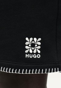 Μαύρο ύφασμα με λευκό λουλουδάτο λογότυπο πάνω από τη λέξη "HUGO" και λευκή ραφή κατά μήκος της άκρης του ρεβέρ.