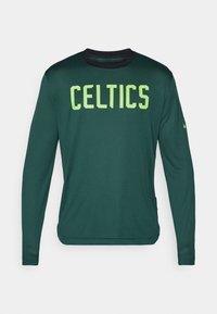 Žalia ilgomis rankovėmis marškiniai su ryškiai geltonais užrašais "CELTICS", juodas apykaklė ir tekstūruota medžiaga, ant rankov�ės yra mažas logotipas.