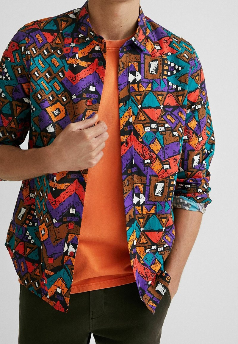 Homme portant une chemise à manches longues à motif géométrique multicolore par-dessus un t-shirt orange, une main ajustant la chemise et l'autre dans la poche.
