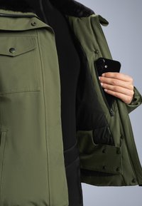 Olive zelena jakna z mehkim črnim podložkom, opremljena z zadrgo z notranjim žepom ter gumbi na sprednjem delu in žepih.