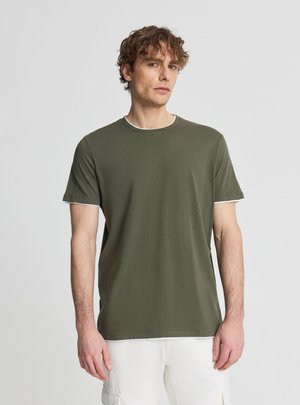 Uomo con capelli ricci che indossa una maglietta verde oliva a maniche corte e pantaloni bianchi, in piedi davanti a uno sfondo chiaro e semplice.