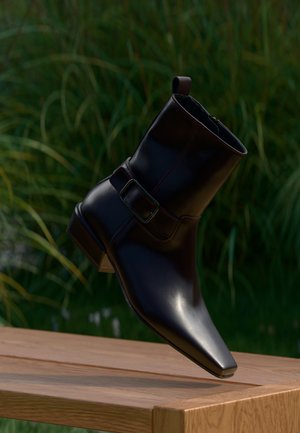 Stivaletto nero in pelle con punta quadrata e fibbia laterale, posizionato sopra una panchina di legno con sfondo di erba verde.