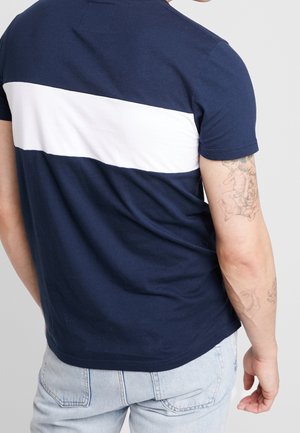 Camiseta azul marino con una franja blanca horizontal en la parte posterior. Manga corta, tejido de algodón y ajuste casual. Tatuaje visible en el brazo.