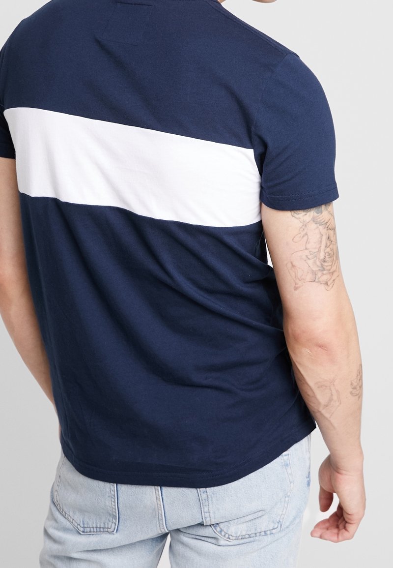 Mörkblå T-shirt med ett horisontellt vitt rand över ryggen. Korta ärmar, bomullstyg och avslappnad passform. Synlig armtatuering.