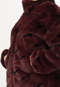 Burgundyfärgad fuskpälsjacka med en mjuk textur, prydd med svarta svish-mönster, hög krage och sidofickor.