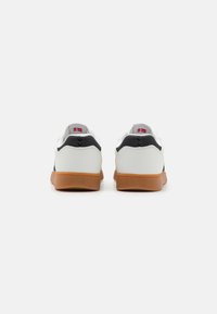 White sneakers featuring a black heel cap and gum rubber outsole. Smooth texture with minimalistic design and subtle logo detail on the heel.