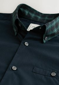 Marineblauw shirt met een groene geblokte kraag, met een knoopsluiting aan de voorkant, een borstzak en plastic knopen. Glad stoftextuur.