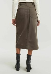 Gonna midi khaki con taglio dritto, tasche posteriori e dettagli di cucitura a vista. Abbinata a stivali neri con tacco.