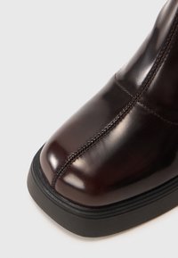 Acercamiento de una bota de cuero marrón oscura pulida con costuras centrales prominentes y suela negra gruesa sobre un fondo claro.