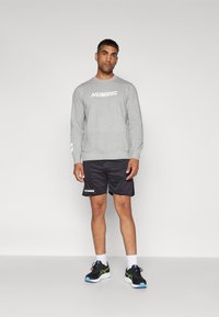 Grijze sweatshirt met "HUMMEL" logo, zwarte shorts met logo, en zwarte sneakers met blauwe en groene accenten. Effen achtergrond, staande pose.
