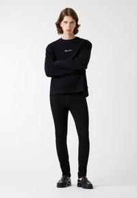 Czarna bluza z okrągłym dekoltem i haftowanym logo, zestawiona z czarnymi, dopasowanymi jeansami i czarnymi butami wsuwanymi, na jednolitym tle.