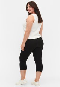 Witte mouwloze top met een geribbelde textuur, gecombineerd met zwarte knielange leggings. De outfit wordt afgemaakt met witte sneakers.