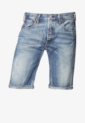 Uomo che indossa una camicia azzurra chiara con motivi e bottoni, pantaloncini di jeans, sneaker bianche e tiene una borsa nera sulla spalla.