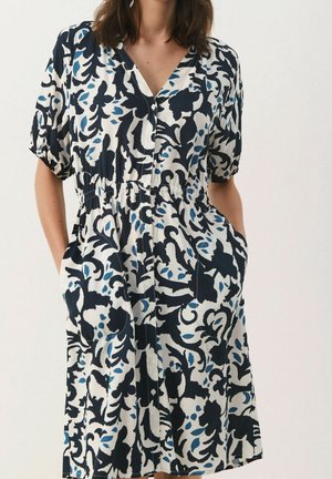 Frau trägt ein geknöpftes Kleid mit kurzen Puffärmeln und einem schwarz-weiß-blauen Blumenmuster, Hände in den Taschen, vor einem schlichten Hintergrund.