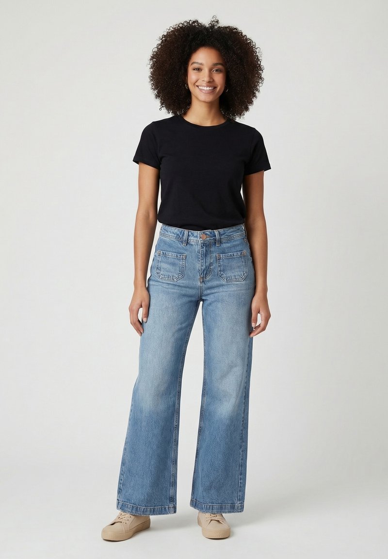 Young woman with curly hair standing, wearing a black t-shirt, wide-leg blue jeans, and beige sneakers, smiling against a plain background.