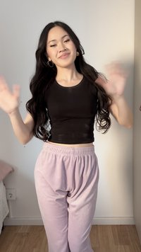 Jeune femme aux longs cheveux foncés portant des boucles d'oreilles créoles dorées, un crop top noir et un pantalon violet clair, faisant signe de la main à l'intérieur.