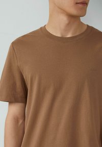 Bruine katoenen T-shirt met een ronde halslijn, korte mouwen en een minimale textuur; heeft een klein logo geborduurd op de borst.