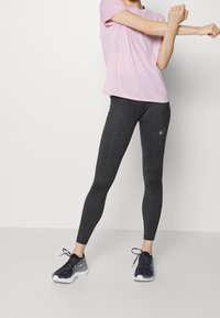 Conjunto deportivo femenino: camiseta de manga corta rosa claro, leggins gris oscuro con logo y zapatillas de deporte negra/gris, en una pose de estiramiento de pie.