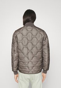 Gevoerde pufferjas in lichtbruin met een rond patroon, ribbelboorden en kraag. Glad oppervlak met een licht glanzende afwerking.