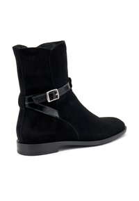 Botín negro de ante hasta el tobillo con una correa de piel negra lisa y detalle de hebilla plateada, que presenta una punta redondeada y suela de goma plana.