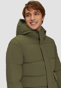 Olivgrüne Steppjacke mit Kapuze, ausgestattet mit einem Reißverschluss, genähten Paneelen und zwei Vordertaschen. Weiches, isoliertes Material.