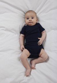 Bebeluș întins pe lenjerie de pat albă, purtând un body albastru închis, privind înainte cu o expresie neutră.