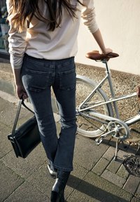 Šedé rozšířené džíny s zadními kapsami, v kombinaci se světle zbarveným topem. Viditelná je černá taška přes rameno a kolo s hnědým sedlem.