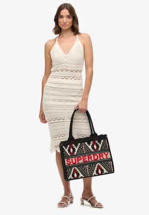 Vestido blanco de punto con textura estampada y dobladillo deshilachado. Bolso tote negro con diseños geométricos y la palabra "SUPERDRY" en rojo.
