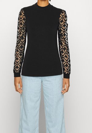 Czarna bluzka z długim rękawem, z przezroczystymi rękawami w wzory i jednolitym przodem, noszona z jasnoniebieskimi szerokimi dżinsami.