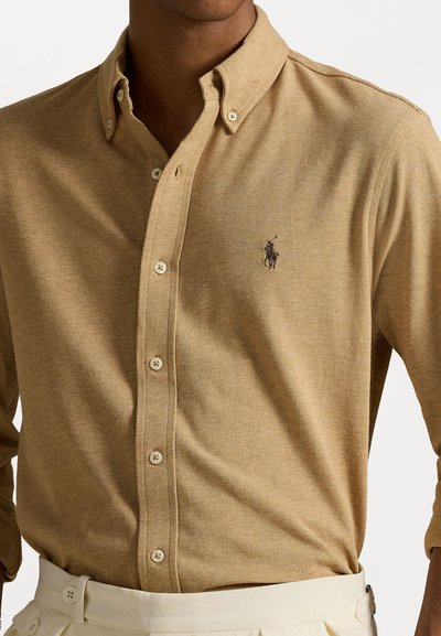 Camisa bege de botões com colarinho estruturado, apresentando um logótipo bordado, tecido texturizado e botões brancos. Estilizada com calças de cor clara.