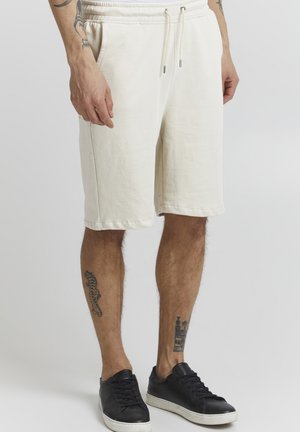 Shorts en coton blancs avec taille à cordon et poches latérales, affichant une coupe décontractée et un design jusqu'aux genoux. Associés à des baskets noires.