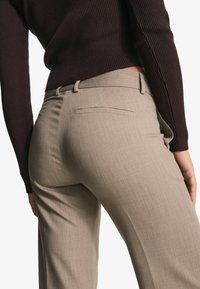 Béžové kalhoty na míru s jemnou texturou látky, s plochým předním dílem, rovnými nohavicemi a jednou zadní kapsou. Kombinovány s tmavým žebrovaným topem.
