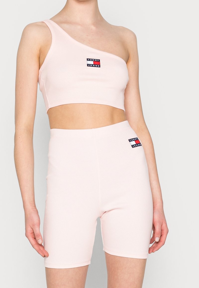 Ljust rosa ribbad crop-top med asymmetrisk design för ena axeln och matchande högmidjade shorts. Har logotyp-patchar på båda plaggen.