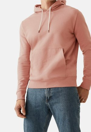 Mand iført en lyserød hættetrøje med hvide snore og forlommer, kombineret med blå denim jeans, stående mod en hvid baggrund.
