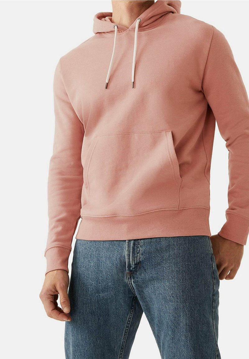 Homme portant un sweat à capuche rose clair avec cordons blancs et poche avant, associé à un jean en denim bleu, debout devant un fond blanc.