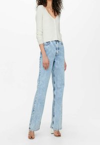 Kobieta w białym, prążkowanym kardiganie zapinanym na guziki, jasnoniebieskich, wysokich dżinsach z szerokimi nogawkami oraz beżowych sandałach na obcasie, stojąca na białym tle.